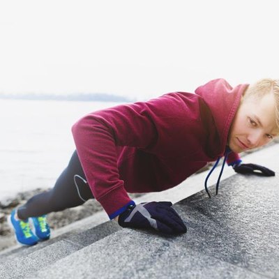 A profile DSLR photo of young male athlete wearing blue running shoes, black tights (pantyhose) and crimson t-shirt with hood. He is doing incline push ups leaning over the stairs of embankment. He is alone in this cold morning. He is looking towards the camera.
