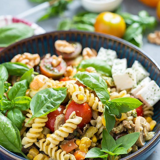 A bowl of colorful pasta salad with bright red tomatoes, green basil leaves, and white chunks of tofu on the side