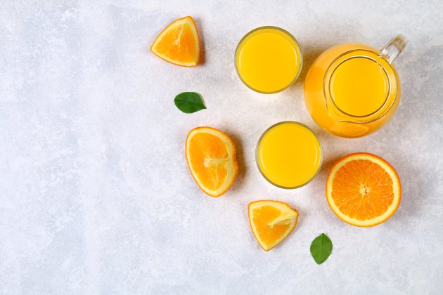 Glass bottles, glasses and a pitcher of fresh orange juice with slices of orange and yellow tubes on a light gray table. Top view. Flat Lay.
