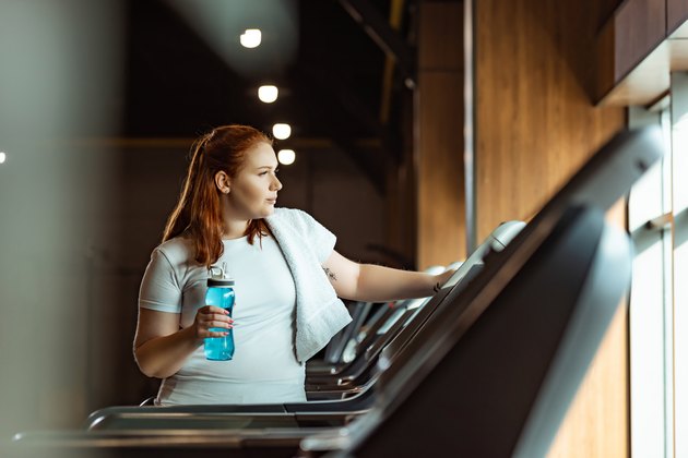 Selective focus of overweight girl with towel on shoulder holding sports bottle while standing at treadmill