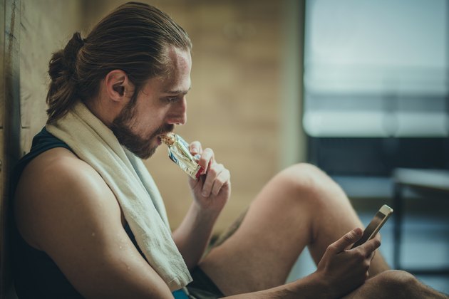 Sportsman eating protein bar and using mobile phone at gym's locker room.