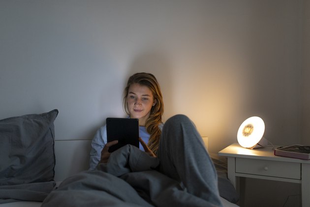Young woman lying in bed at home at night using tablet