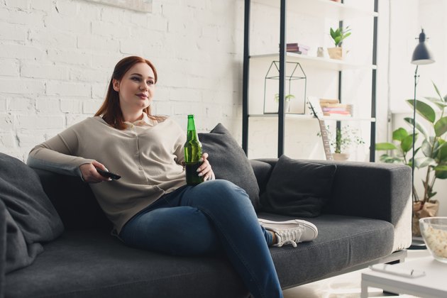 beautiful woman holding bottle of beer and remote control at home