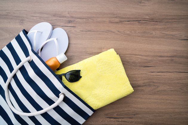 white flip flops yellow towel and black sunglasses on wooden floor coming out of striped beach bag