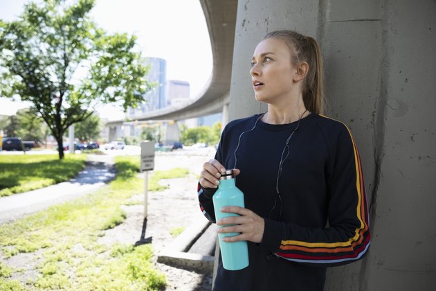 Young female runner with water bottle resting under urban overpass