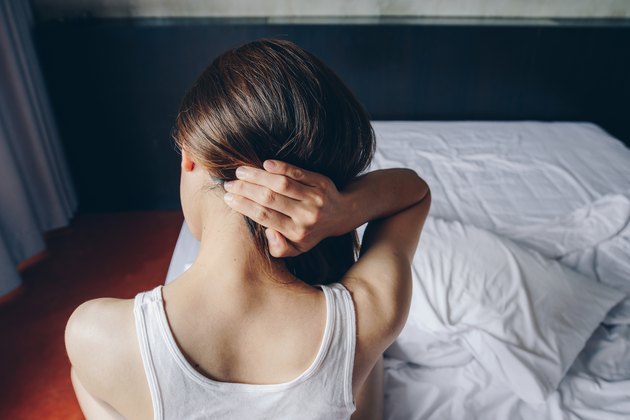 Back view of young woman sitting on bed holding the back of her neck