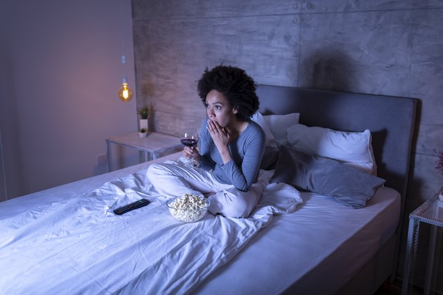 A woman watching a movie on her bed while eating popcorn and drinking a glass of wine