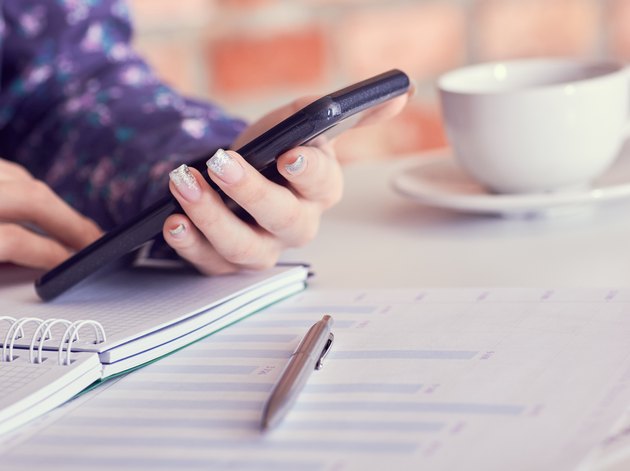 Close-up of female hands using smart phone while working on computer at modern office interior, businesswoman typing text message on her cellphone at loft interior.