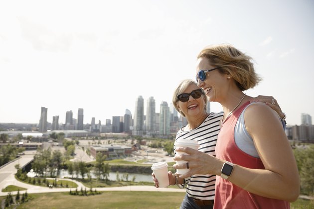 Smiling mother and daughter walking in sunny urban park with coffee