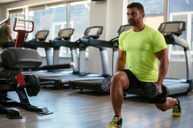 photo of man in gym in front of treadmills doing lunges holding dumbbells