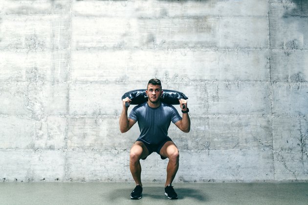 Caucasian bodybuilder with serious facial expression working out with Bulgarian Training bag. In background wall.