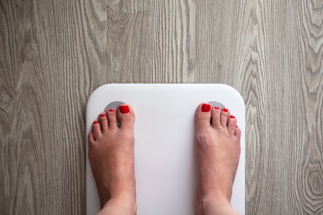 Woman stands on white modern electronic sensor scales. Only feet are visible. Scales stand on gray wooden floor. Space for text. Healthy lifestyle, diet, weight loss concept. Horizontal.