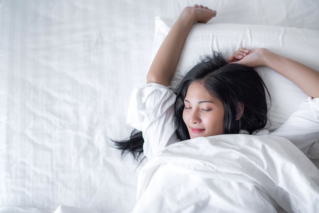 美丽的年轻漂亮的亚洲女人醒来，让快乐的微笑与白衬衫在白色的床在早上。