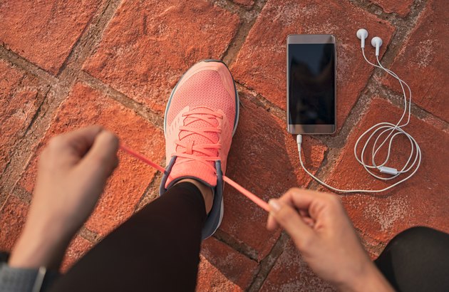 女跑步者系好鞋带准备在跑道上比赛，用智能手机和耳机听手机上的音乐。运动员为有氧训练做准备。脚在地面。