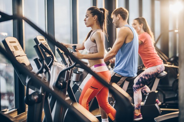 Three athletic people warming up on exercise machines in a gym.