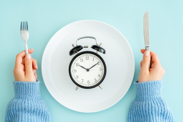 A woman holding a fork and knife next to a plate with an alarm clock to symbolize intermittent fasting
