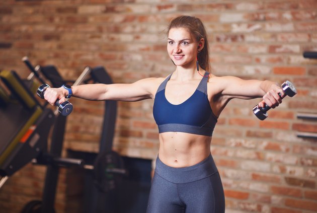 Portrait of attractive cheerful sportswoman with headphones in her ears. Sporty woman doing exercises with dumbbells for shoulders in the gym on the background of brick wall