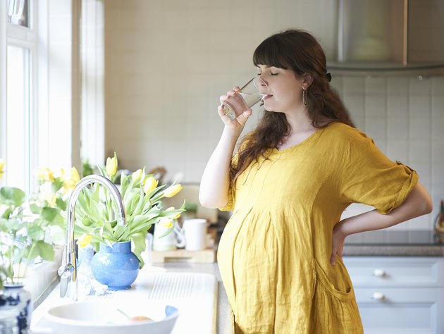 Pregnant woman drinking glass of water in kitchen at home.