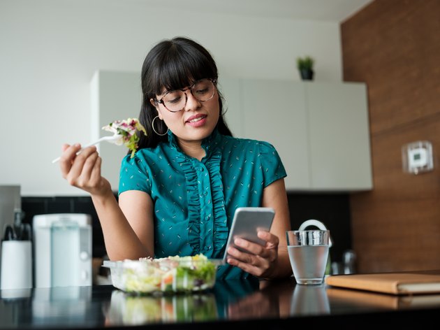 Young businesswoman eating salad in lunch room and using phone