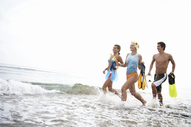 Three friends running in water on beach, holding flippers, smiling