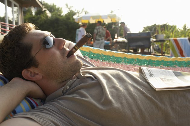 Man lying in hammock smoking cigar, friends by barbecue in background