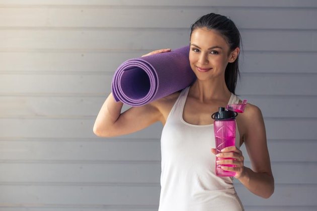Beautiful young woman in sports wear is holding a yoga mat and a bottle of water, looking at camera and smiling, standing on a gray background