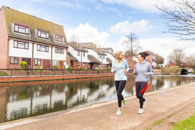 Two girls jogging outdoors in London. They are caucasian women, looking each other while running along a canal with some beautiful houses on background on a sunny day in spring.