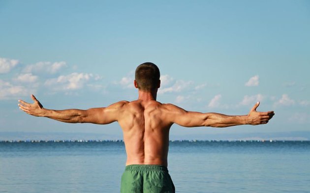 Man Raising His Hands or Open arms, looking to sea blue sky horizont. Strong muscular man, perfect body, arms, back. Freedom Concept