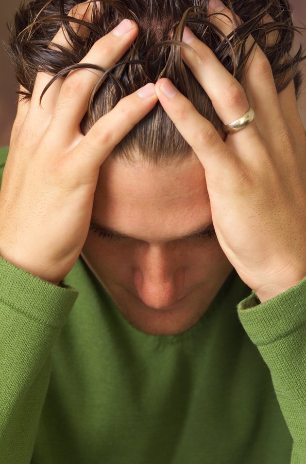 caucasian young man wearing green shirt looks down as he holds his head in hands fingers in hair