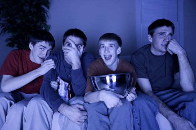 Four teenage boys (13-14) (16-17) sitting on couch watching TV, eating snacks