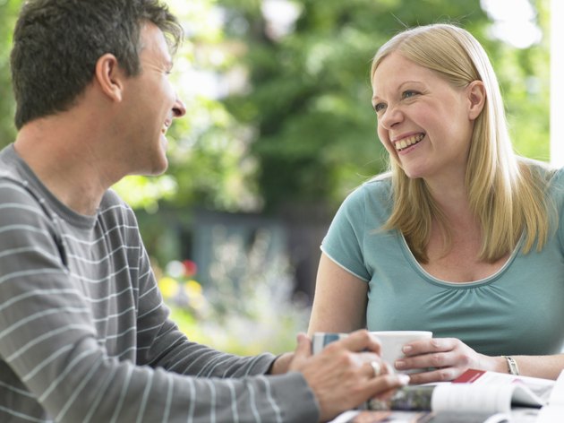 Couple sitting at table holding mugs, looking at each other, laughing