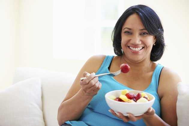 Overweight Woman Sitting On Sofa Eating Bowl Of Fresh Fruit