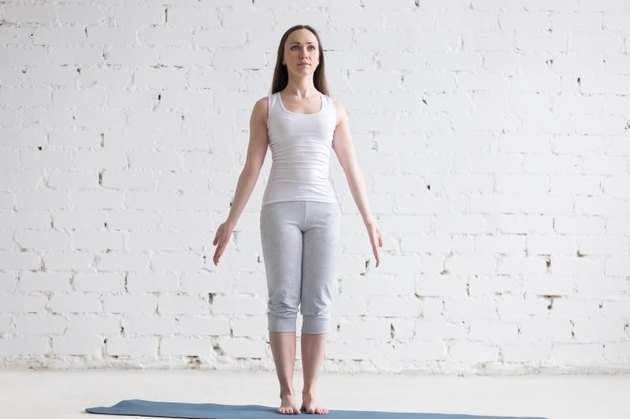 Attractive happy young woman working out indoors. Front view portrait of beautiful model doing yoga exercise on blue mat. Standing in Tadasana, Mountain Pose. Full length