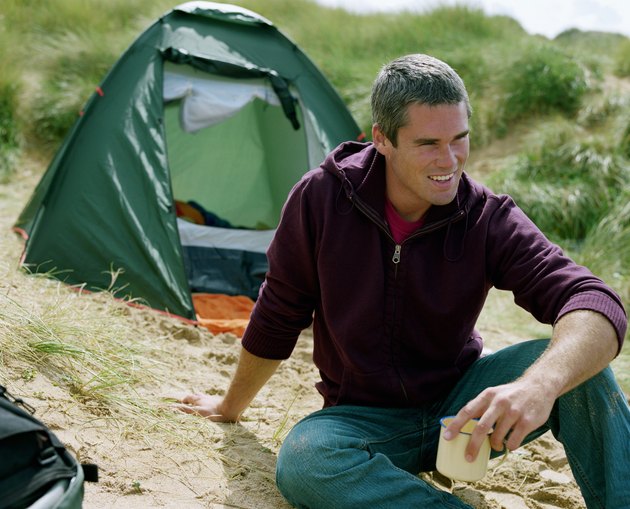 Young man sitting on sand by tent, holding cup, looking away, smiling