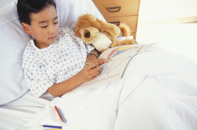 Boy (6-7) lying in hospital bed with toy animal, drawing in book