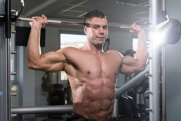 Muscular Young Man Doing Heavy Weight Exercise For Shoulders In Gym