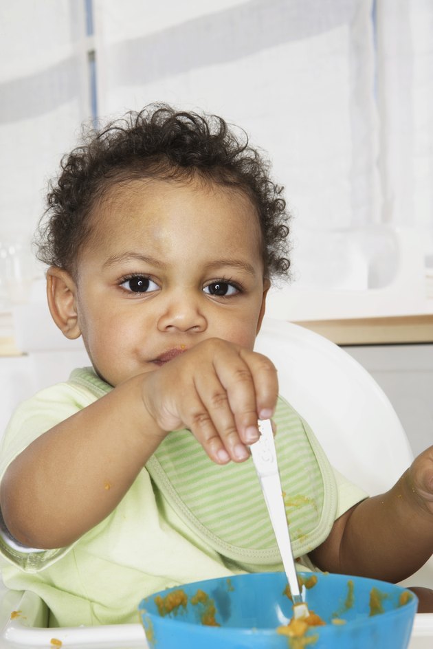 Baby boy (18-24 months) in high chair holding spoon in bowl, portrait