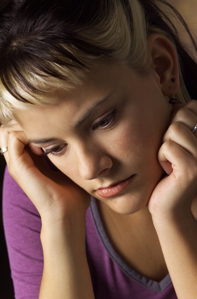 a caucasian young woman with brown and blond streaked hair wearing a purple shirt is looking down sadly as she leans on her hands