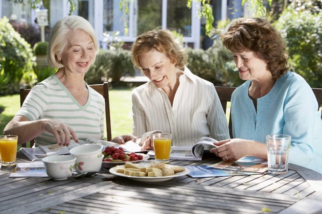 Three senior women in garden looking at holiday brochures, smiling