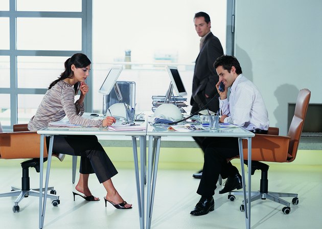 Two Business Colleagues Working Face to Face at Their Desks With Another Businessman Walking Past in the Office