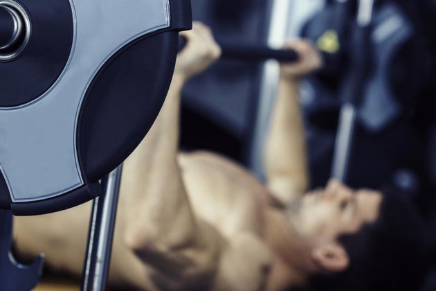 Man lifting weights, lying on bench press in gym. Focus on weight plate, model blurred. Color graded image, convenient copy space.