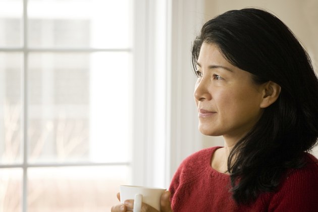 Pensive mature Asian woman at window holding cup of tea