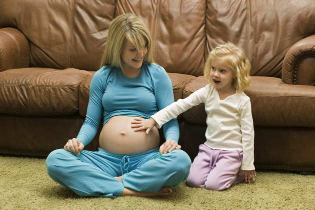 Daughter (4-5) touching belly of pregnant mother sitting on floor by sofa