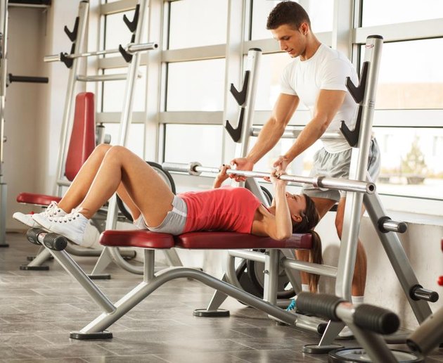 Attractive woman lies on a bench and performs barbell weights presses at the fitness center.Her fitness instructor assists.