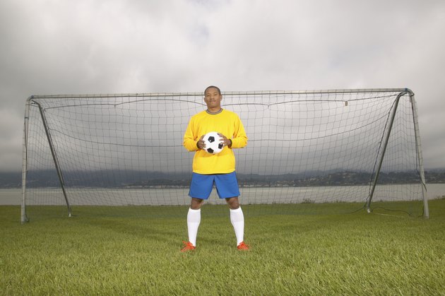 Young male football player standing at a goal post holding a soccer ball