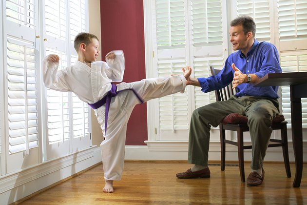 A boy in a martial arts uniform practices a side kick into his father's hand