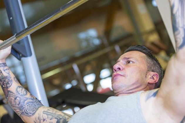 A muscular, tattooed man performs and incline barbell bench press at the gym.