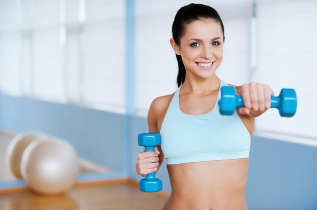 Beautiful young woman in sports clothing exercising with dumbbells and smiling while standing in health club