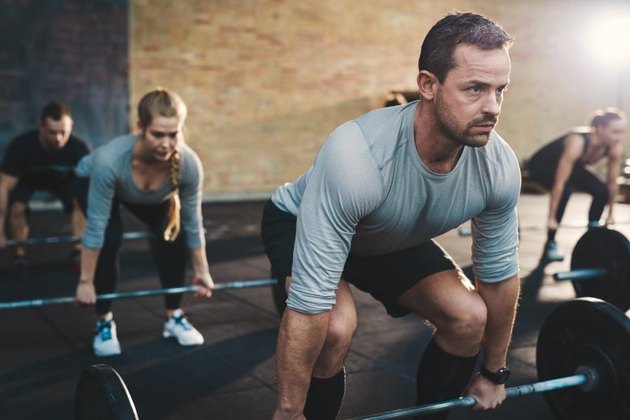 Fit young man lifting barbells looking focused, working out in a gym with other people