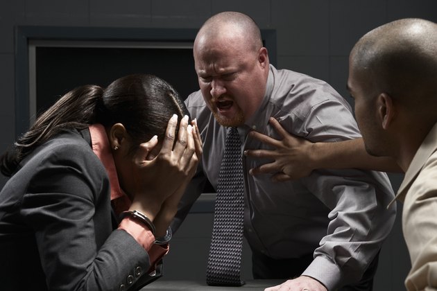 Two men and handcuffed woman at desk in interrogation room, man shouting at woman